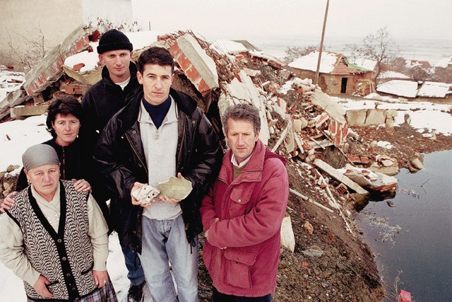 Kosovska družina ob kraterju, kjer je nekoč stala njihova hiša, ki jo je pomotoma uničila Natova bomba. Vas stoji tik nad vojaškim delom prištinskega letališča Slatina. Ostanke bombe so domačini shranili, da bi tako morda lažje prišli do nekakšne odškodnine za porušen dom.