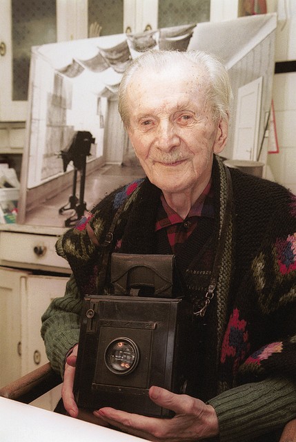Peter Lampič (maja bo praznoval 102. leto) najstarejši slovenski fotograf v kuhinji svoje hiše (maketa hiše, ki jo je naredil sam, stoji pred njim). Zadaj je slika ateljeja, kjer je trenutno skladišče.
