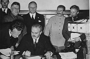 Molotov podpisuje dokument; za njem stojita Ribbentrop in Stalin