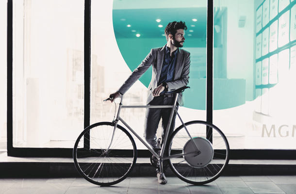 Bicikel s FlyKly Smart Wheelom, ki ga prepoznamo po belem kolutu na zadnjem kolesu. Ob njem stoji podpornik FlyKlyeve kampanje, industrijski oblikovalec Jurij Lozić, ki je pred časom na Kickstasterju tudi sam prepričal z avtorskim projektom, namenjenim kolesarjem.