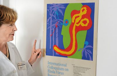 Na notranji strani vhodnih vrat pisarne visi plakat mednarodne konference o eboli, ki se je je udeležila leta 1996