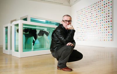 Damien Hirst pred svojima umetninama: na levi je delo Mother and Child (Divided) (Prepolovljena mati in otrok), s katerim se je leta 1993 predstavil na Beneškem bienalu, na desni ena od slik z barvnimi pikami.