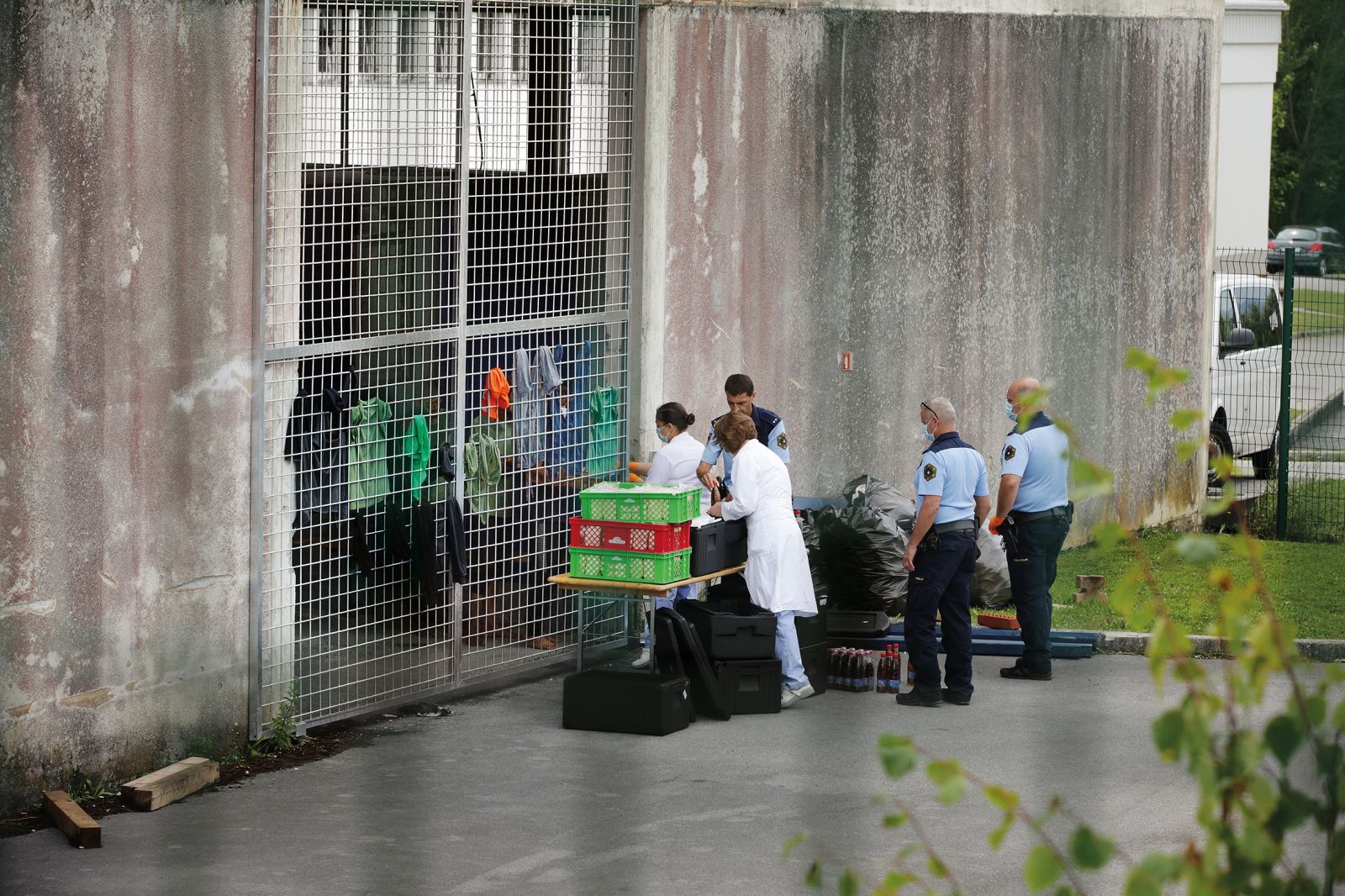 Realno stanje: čas kosila za zaprte v Centru za tujce v Postojni. Nekateri dobijo hrano skozi nekaj centimetrsko režo v mreži, s katero so zaprti v opuščenem industrijskem objektu. 