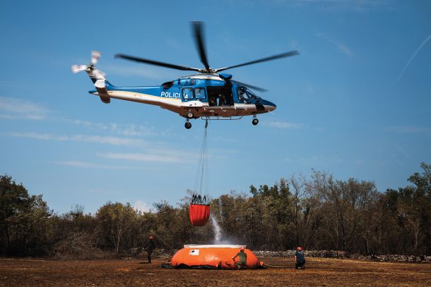 Slovenski policijski helikopter med zajemanjem vode blizu Opatjega sela. Helikopter polni podvesno vrečo z 800 litri vode. Hrvaški kanader jih v enem zamahu odvrže 6200. 