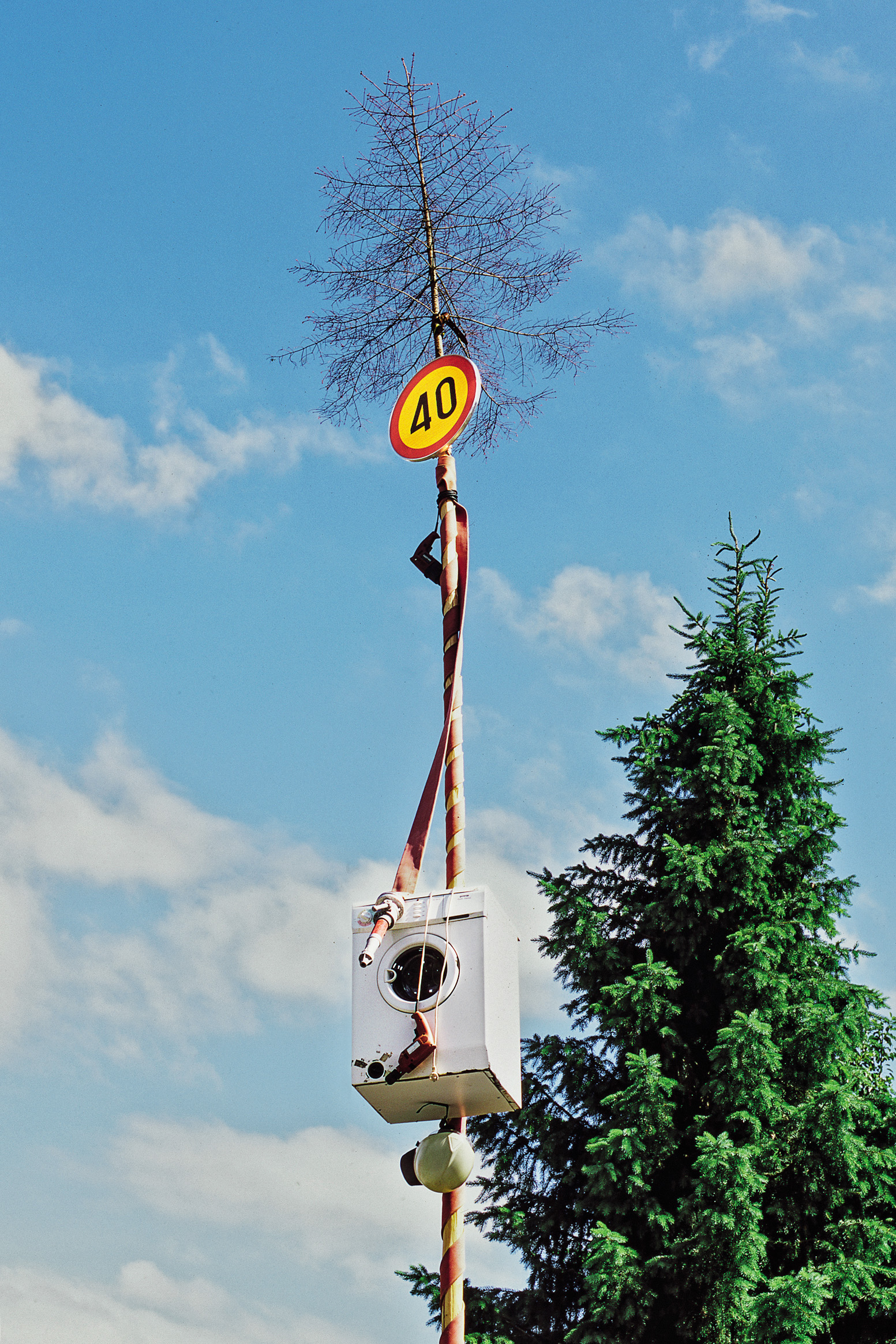 Rojstnodnevni mlaj, okrašen s pralnim strojem, gasilsko cevjo in bormašino. Kaj točno nam nenavadna kombinacija predmetov sporoča, je stvar interpretacije.